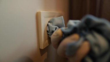 Bir temizlikçinin eli, beyaz bir duvara mavi bir soketi dikkatlice siler, yakın plan. Profesyonel temizlik. Temiz ev, antibakteriyel temizlik, toz alma. Elektrik ve güvenlik kuralları.