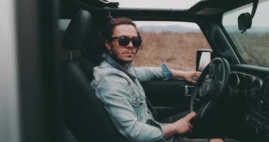 Genç bir adam direksiyonun arkasında güneş gözlüklü ve kot ceketli bir arabada oturuyor.