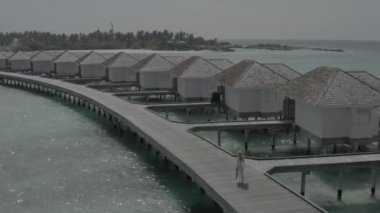 Beyaz elbiseli bir kız, bir otel bungalovunun yakınındaki ahşap bir köprü boyunca yürüyor. Maldivian adası tatil köyü. Maldivler. Cennette yaz tatili. Renk düzeltmesi olmayan görüntü.