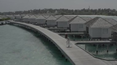 Beyaz elbiseli bir kız, bir otel bungalovunun yakınındaki ahşap bir köprü boyunca yürüyor. Maldivian adası tatil köyü. Maldivler. Cennette yaz tatili. Renk düzeltmesi olmayan görüntü.