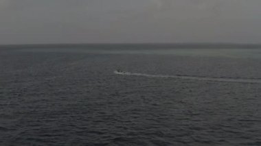 Açık bir yaz gününde arka planda küçük bir adayla birlikte Maldivler 'deki Hint Okyanusu' nda yavaşça ilerleyen küçük beyaz bir teknenin hava görüntüsü. Renk düzeltmesi olmayan görüntü.