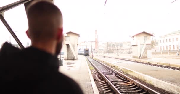 若い男が駅を出る電車を見ている 駅を出発する列車を見ている若者の肖像画 — ストック動画