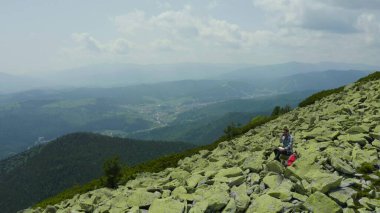Bir adam kayalık bir dağın tepesinde otururken dizüstü bilgisayarda çalışıyor. Dağlarda tatildeyken uzaktan kumandalı iş. Dünyanın herhangi bir yerinde uzaktan çalışma kavramı.