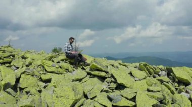 Bir adam kayalık bir dağın tepesinde otururken dizüstü bilgisayarda çalışıyor. Dağlarda tatildeyken uzaktan kumandalı iş. Dünyanın herhangi bir yerinde uzaktan çalışma kavramı.