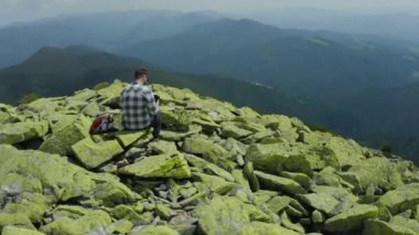 Bir adam kayalık bir dağın tepesinde otururken dizüstü bilgisayarda çalışıyor. Dağlarda tatildeyken uzaktan kumandalı iş. Dünyanın herhangi bir yerinde uzaktan çalışma kavramı.