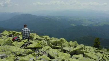 Bir adam kayalık bir dağın tepesinde otururken dizüstü bilgisayarda çalışıyor. Dağlarda tatildeyken uzaktan kumandalı iş. Dünyanın herhangi bir yerinde uzaktan çalışma kavramı.