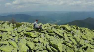 Bir adam kayalık bir dağın tepesinde otururken dizüstü bilgisayarda çalışıyor. Dağlarda tatildeyken uzaktan kumandalı iş. Dünyanın herhangi bir yerinde uzaktan çalışma kavramı.