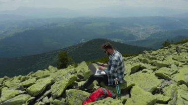 Bir adam kayalık bir dağın tepesinde otururken dizüstü bilgisayarda çalışıyor. Dağlarda tatildeyken uzaktan kumandalı iş. Dünyanın herhangi bir yerinde uzaktan çalışma kavramı.