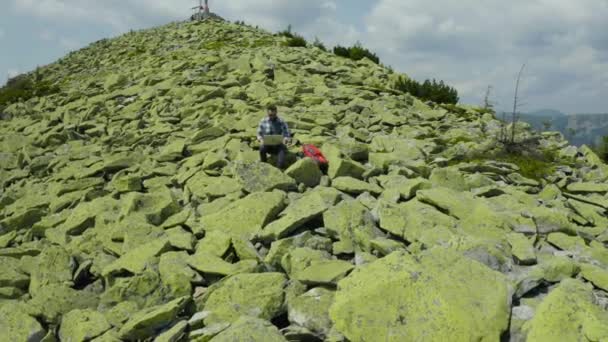一个男人坐在一座岩石山的顶上 一边在笔记本电脑上工作 在山里度假时做远程工作 世界任何地方的远程工作的概念 — 图库视频影像