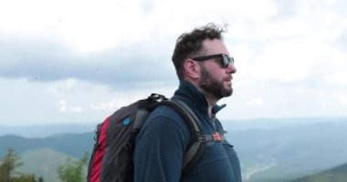Tırmanan bir adamın portresi. Sırt çantalı bir turist, dağlarda yürüyüş yapıyor, etrafa bakıyor. Macera turizmi. Renk düzeltmesi olmadan 4k.
