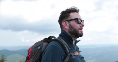 Tırmanan bir adamın portresi. Sırt çantalı bir turist, dağlarda yürüyüş yapıyor, etrafa bakıyor. Macera turizmi. Renk düzeltmesi olmadan 4k.