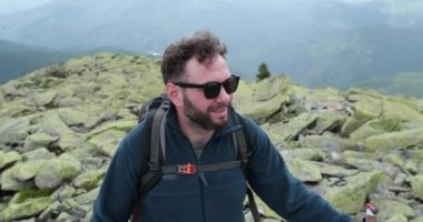 Tırmanan bir adamın portresi. Sırt çantalı bir turist, dağlarda yürüyüş yapıyor, etrafa bakıyor. Macera turizmi. Renk düzeltmesi olmadan 4k.