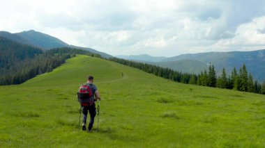 Sırtında sırt çantasıyla dağlarda bir adam güzel bir yaz gününde çimenli bir yamaç boyunca yürür. Dağ yürüyüşü kavramı. Dağlarda tek başına turist gezintisi.