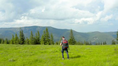 Sırtında sırt çantasıyla dağlarda bir adam güzel bir yaz gününde çimenli bir yamaç boyunca yürür. Dağ yürüyüşü kavramı. Dağlarda tek başına turist gezintisi.