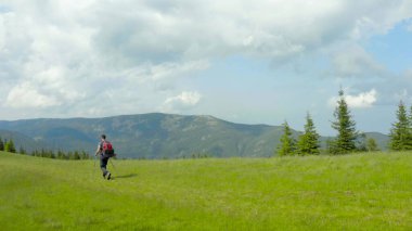 Sırtında sırt çantasıyla dağlarda bir adam güzel bir yaz gününde çimenli bir yamaç boyunca yürür. Dağ yürüyüşü kavramı. Dağlarda tek başına turist gezintisi.