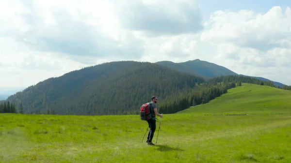 背中のバックパックを持った山の男は 美しい夏の日に草の斜面に沿って歩きます 山岳ハイキングのコンセプト 山の単一の観光ハイキング — ストック写真