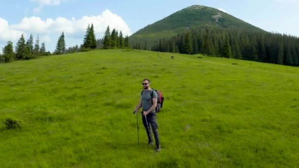 背中のバックパックを持った山の男は 美しい夏の日に草の斜面に沿って歩きます 山岳ハイキングのコンセプト 山の単一の観光ハイキング — ストック動画