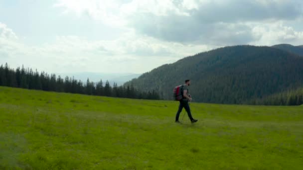 在一个美丽的夏日里 一个背着背包的山区男子沿着草坡散步 登山的概念 一个游客在山上徒步旅行 — 图库视频影像