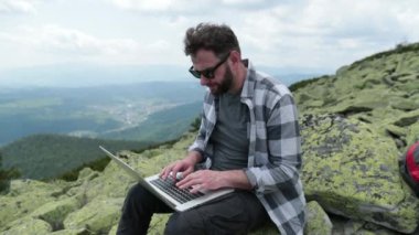 Dağın tepesinde dizüstü bilgisayarla çalışan genç bir gezgin. Dünyanın herhangi bir yerindeki uzaktan çalışma kavramı..