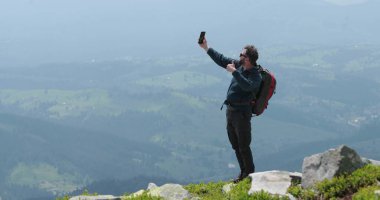 Dağın tepesindeki bir gezgin telefonla konuşuyor, fotoğraf çekiyor ve görüntülü konuşma yapıyor. Dünyanın uzak yerlerinde iletişim kavramı.