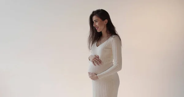 美丽的怀孕母亲穿着白色背景的衣服在工作室里摆姿势 一位身穿白衣的年轻的黑发孕妇正在抚摸着她的腹部 享受着怀孕的快乐 — 图库照片