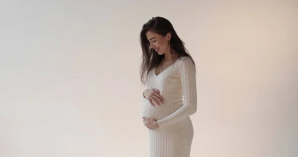 Güzel hamile anne beyaz arka planda stüdyoda poz veriyor. Beyaz elbiseli, genç, esmer bir kadın karnını okşuyor ve hamileliğinin tadını çıkarıyor..