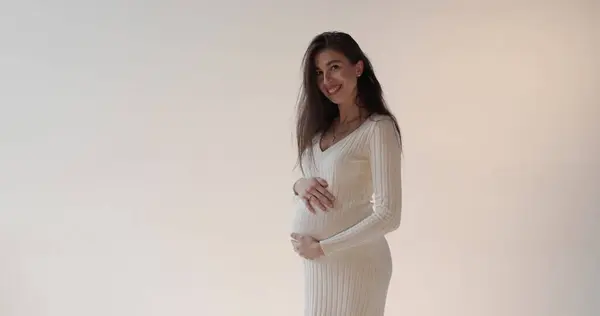 白い背景にスタジオでポーズする美しい妊娠中の母親 白いドレスを着た若いブルネットの妊婦が彼女の腹を打って妊娠期間を楽しんでいる — ストック写真
