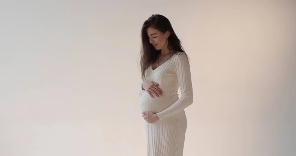 Güzel hamile anne beyaz arka planda stüdyoda poz veriyor. Beyaz elbiseli, genç, esmer bir kadın karnını okşuyor ve hamileliğinin tadını çıkarıyor..