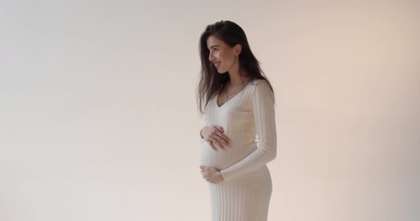 白い背景にスタジオでポーズする美しい妊娠中の母親 白いドレスを着た若いブルネットの妊婦が彼女の腹を打って妊娠期間を楽しんでいる — ストック動画
