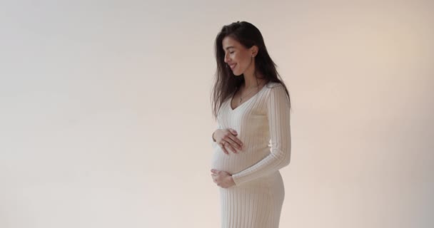 白い背景にスタジオでポーズする美しい妊娠中の母親 白いドレスを着た若いブルネットの妊婦が彼女の腹を打って妊娠期間を楽しんでいる — ストック動画
