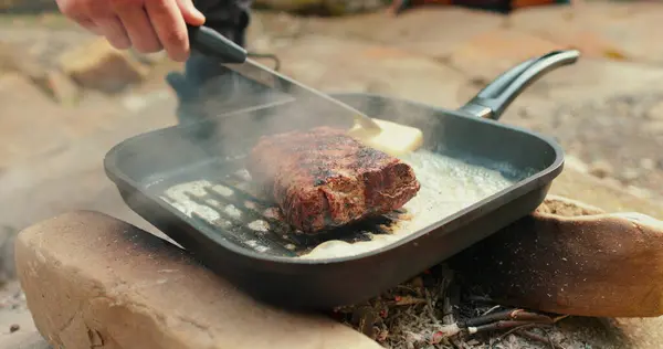 Adding butter to a steak on a hot pan in the process of grilling meat outdoors. Cooking meat on an open fire in nature on a tourist trip.