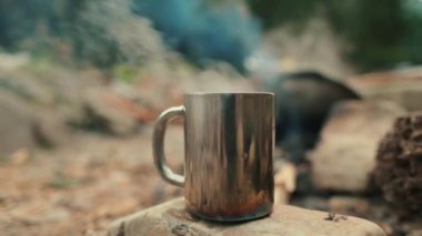 Şenlik ateşinin arkasındaki taşın üstünde metal bir turist kupası. Kamp gezisinde sıcak içeceği olan bir kupa..