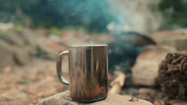 Şenlik ateşinin arkasındaki taşın üstünde metal bir turist kupası. Kamp gezisinde sıcak içeceği olan bir kupa..