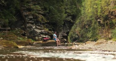 Gezgin bir adam yaz ormanındaki bir nehrin kenarındaki bir taşın üzerinde oturarak su içer. Sırt çantalı bir erkek turist dağlarda yürüyüş yaparken nehir kenarında dinleniyor..