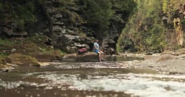 Gezgin bir adam yaz ormanındaki bir nehrin kenarındaki bir taşın üzerinde oturarak su içer. Sırt çantalı bir erkek turist dağlarda yürüyüş yaparken nehir kenarında dinleniyor..