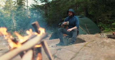 Bir gezgin, dağlarda bir turist yürüyüşü sırasında kamp ateşinin yanındaki metal bardaktan çay içer.