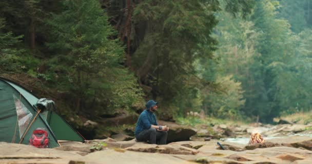 孤独な旅行者は川岸のキャンプファイヤーの近くの観光キャンプにいる 川岸の旅行者のキャンプ — ストック動画