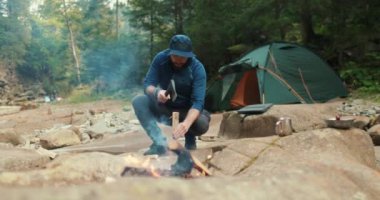 Erkek bir turist ormanda kamp ateşinin yanında küçük bir baltayla odun kırıyor. Gezgin ateş için odun hazırlar. Vahşi doğadaki adam. Doğa yürüyüşçüsü, vahşi yaşam, kamp ateşi.
