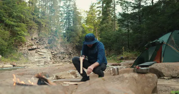 Erkek bir turist ormanda kamp ateşinin yanında küçük bir baltayla odun kırıyor. Gezgin ateş için odun hazırlar. Vahşi doğadaki adam. Doğa yürüyüşçüsü, vahşi yaşam, kamp ateşi.