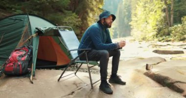 Bir adam dinleniyor, yiyor ve içiyor dağlarda bir çadırın yakınında, bir sandalyede oturuyor. Doğa yürüyüşü. Çadır kampı. Kampta çadırın yanında oturan bir gezgin..