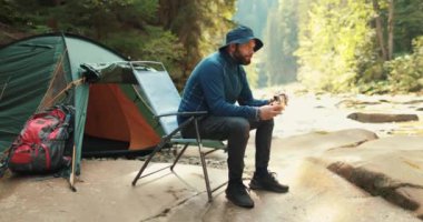 Bir adam dinleniyor, yiyor ve içiyor dağlarda bir çadırın yakınında, bir sandalyede oturuyor. Doğa yürüyüşü. Çadır kampı. Kampta çadırın yanında oturan bir gezgin..