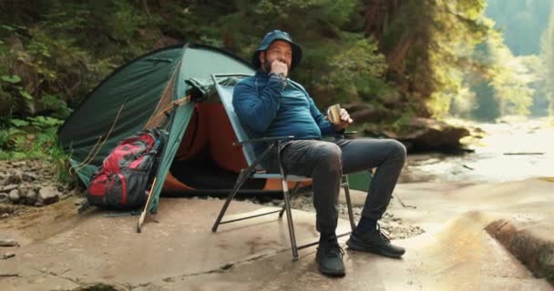 男は休憩し 山のテントの近くで食べ 椅子に座っている 自然の中でハイキング テントキャンプ キャンプのテントの近くに座っている旅行者 — ストック動画