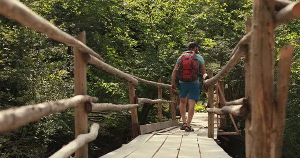 A man in hiking gear with a backpack walks across an epic suspension bridge in a national park. A cinematic adventure of traveling in the mountains. A man walks through a small town on a mountain river.