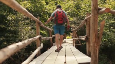 Sırt çantalı yürüyüş kıyafeti giymiş bir adam ulusal parktaki destansı asma köprüden geçiyor. Dağlarda gezmenin sinematik bir macerası. Bir adam dağ nehri üzerinde küçük bir kasabada yürüyor..