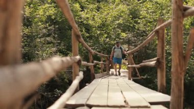 Sırt çantalı yürüyüş kıyafeti giymiş bir adam ulusal parktaki destansı asma köprüden geçiyor. Dağlarda gezmenin sinematik bir macerası. Bir adam dağ nehri üzerinde küçük bir kasabada yürüyor..