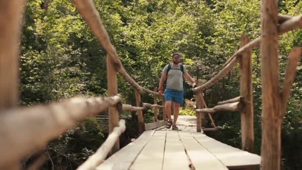 一个背着背包 穿着远足装备的男人穿过国家公园里一座宏伟的悬索桥 在山上旅行的电影冒险 一个人走在山河边的一个小镇上 — 图库视频影像