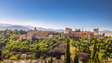 İspanya 'nın Granada kasabasındaki Mirador de San Nicolas' tan Alhambra Panoraması. Bereketli yeşillik ve açık mavi gökyüzünün altındaki dağlarla çevrili bir tepenin üzerine kurulmuş antik saray..