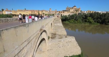 Cordoba, İspanya - 7 Eylül 2024: Durgun bir nehir ve açık mavi gökyüzü altındaki antik binalara bakan Roma köprüsünde turistler geziniyor.