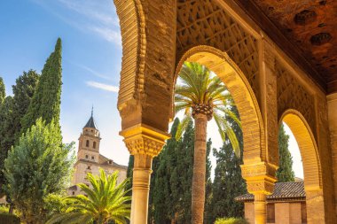 Granada kasabasındaki Alhambra kompleksi, Endülüs, İspanya. Süslü kemerler güneşten sırılsıklam olmuş bir avluyu çerçeveler, palmiye ağaçları ve uzak bir kuleyi ortaya çıkarır..