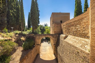 Acequia Real 'in su kemeri Alhambra kompleksi, Granada kasabası, Endülüs, İspanya. Antik taş kemer yolu ve açık mavi gökyüzünün altındaki kale duvarları. Uzun selvi ağaçları sahneyi çerçeveliyor.